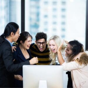 Group of colleagues hugging smiling and excited facing a computer on a desk against a window background