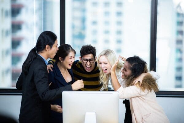 Group of colleagues hugging smiling and excited facing a computer on a desk against a window background