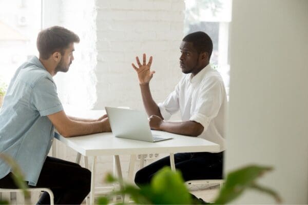 casual dress colleagues on opposite sides of table one with laptop arm raised and other sitting forward listening