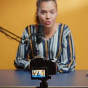 yellow background woman sitting at grey desk talking into mic whilst facing camera with laptop on desk recording herself
