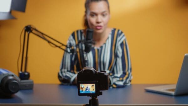 yellow background woman sitting at grey desk talking into mic whilst facing camera with laptop on desk recording herself