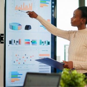 office meeting woman standing at dashboard pointing to graph with pen whilst holding a clipboard whilst colleagues look on