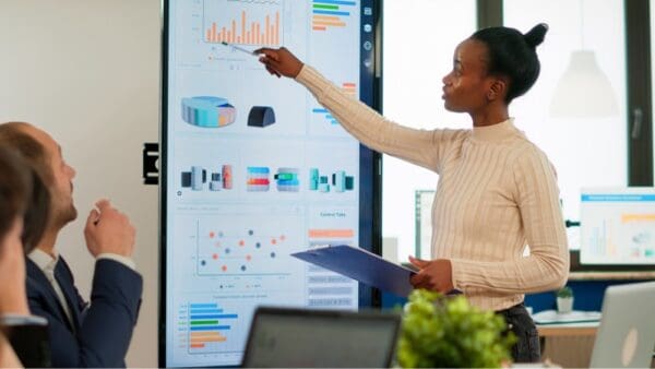 office meeting woman standing at dashboard pointing to graph with pen whilst holding a clipboard whilst colleagues look on